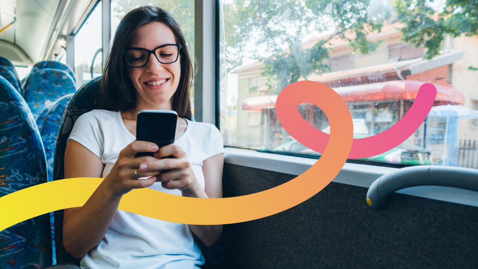 Foto de uma jovem branca, de cabelos lisos e óculos de grau sentada em um banco de ônibus olhando para o celular e sorrindo. Ao lado dela, uma mochila cinza.