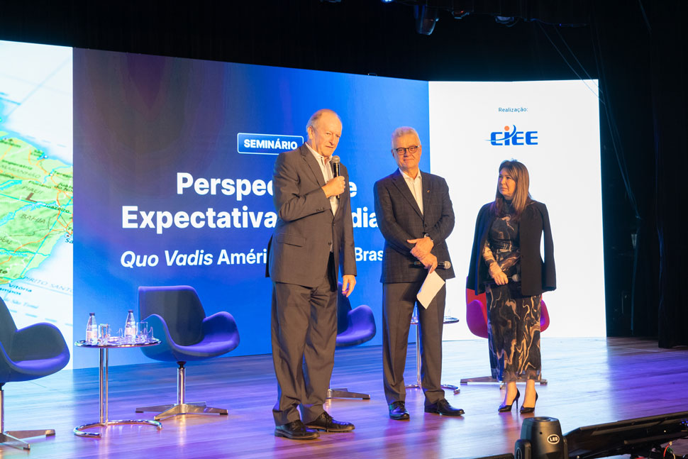 Dois homens à esquerda do palco e uma mulher à direita, todos de pé com vestes sociais, a iluminação é azul e roxa e o painel atrás das pessoas mostra o nome do evento