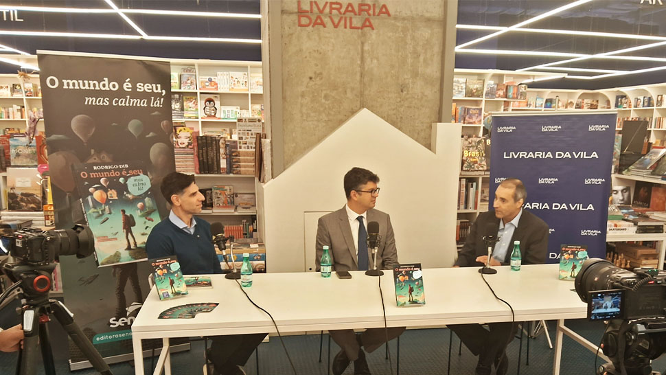 Três homens estão sentados em uma mesa branca, em frente a microfones, participando de uma conversa gravada com câmeras. Ao fundo, estantes com livros e banners do livro “O mundo é seu, mas calma lá” e da Livraria da Vila.
