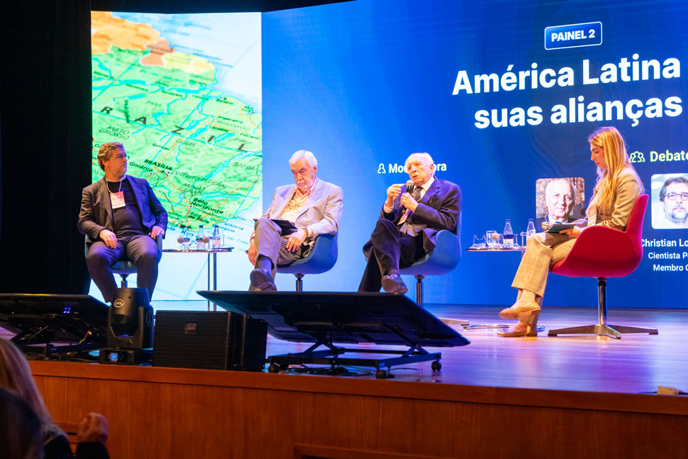 Quatro pessoas sentadas em bancos em cima do palco, com iluminação roxa e azul, uma mulher loira à direita, vestida de blazer bege, comanda o debate.