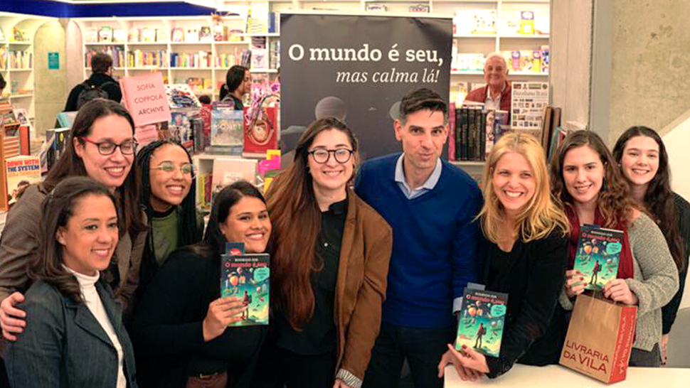 Um grupo de oito mulheres está reunido em volta do autor Rodrigo Dib, todos sorrindo para a foto. Algumas seguram o livro “O mundo é seu, mas calma lá”. Ao fundo, prateleiras de livros e um banner com o nome da obra. A foto foi tirada na Livraria da Vila.