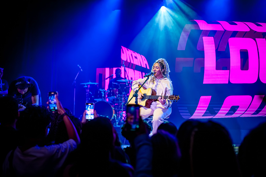 Uma mulher de cabelos cacheados se encontra em cima de um palco, sentada em um banco, com um violão no colo. Ela canta para a plateia.