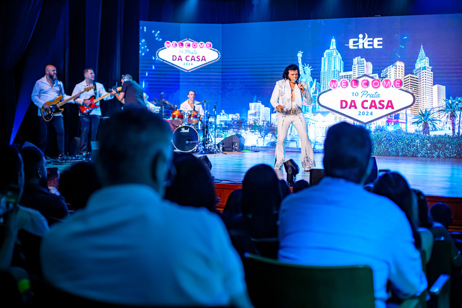 Homem com cabelos médios e escuros, pele branca e roupas brancas. Ele se apresenta em cima do palco do Espaço CIEE