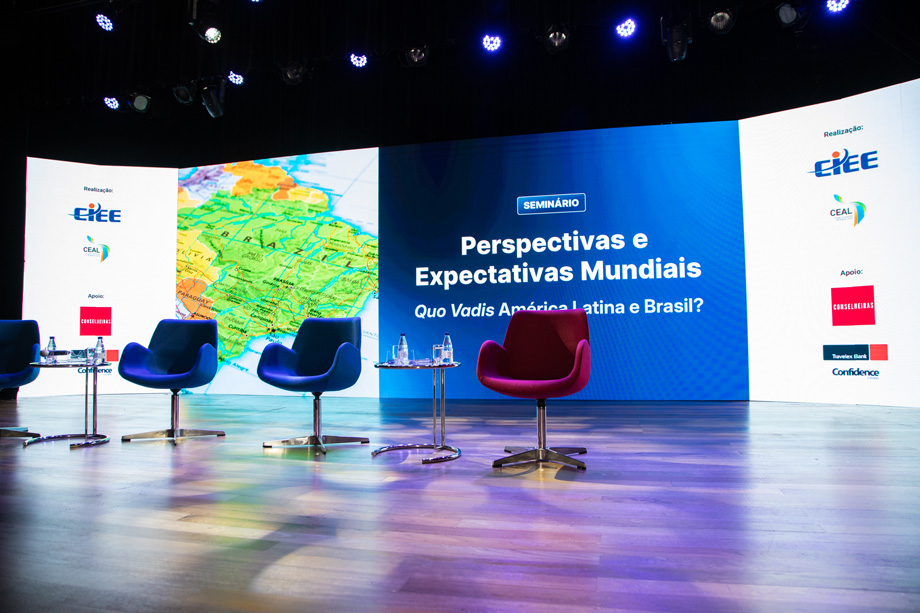 Um palco bem iluminado, do ESPAÇO CIEE, com 4 cadeiras uma ao lado da outra com um grande painel ao fundo