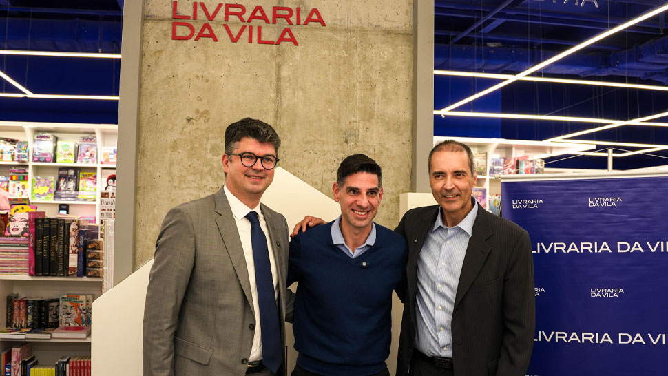 Três homens posam sorrindo para foto em frente a uma coluna com o letreiro “Livraria da Vila”.