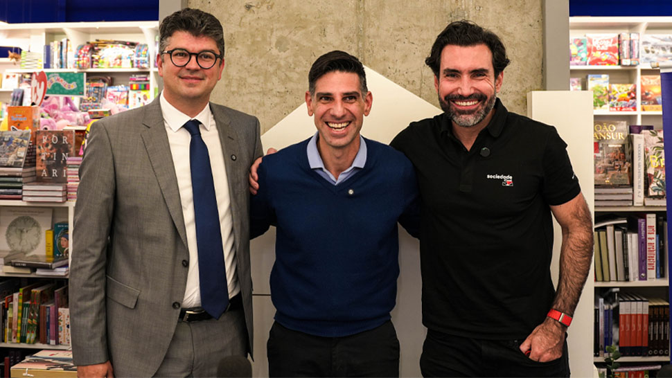 📸 Imagem 4: Três homens, sendo um deles de camisa preta e outros dois em trajes formais, sorriem em frente a uma prateleira de livros.