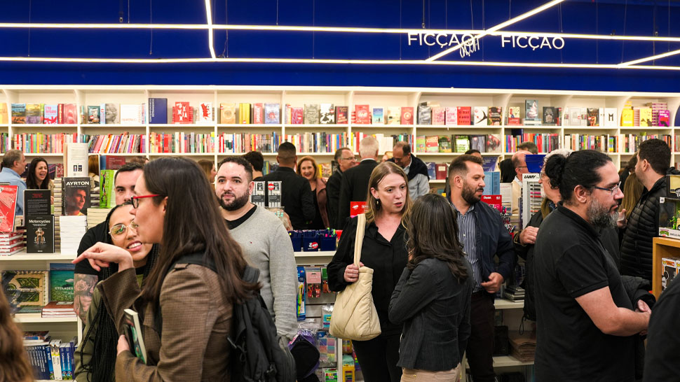 : Livraria cheia de pessoas conversando entre as estantes. O espaço está iluminado e com prateleiras repletas de livros na seção de ficção.
