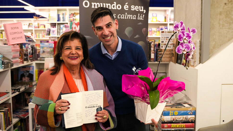 Rodrigo Dib, ao lado de Luiza Helena Trajano, ambo estão em uma biblioteca. Dib segura uma flor na mão e Luiza o livro.