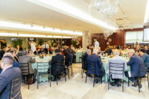 Foto mostra o Foyer cheio de pessoas, sentadas em mesas circulares, há garçons andando pelo local