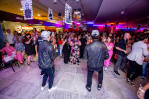 Dois homens estão de jaqueta de couro preta e boné de frente para um publico dançante