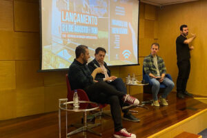 Três homens sentados em um palco do auditório com um painel ao fundo.