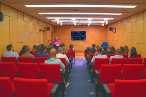 Auditório com paredes amarelas e cadeiras vermelhas. Há algumas pessoas sentadas