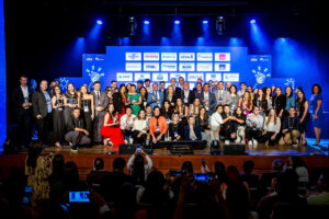 Diversas pessoas, segurando prêmios na mão, em pé no grande palco do teatro com um telão ao fundo