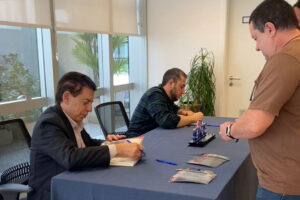 Mostra dois homens com ternos, sentados em uma mesa de recepção, cadastrando os convidados