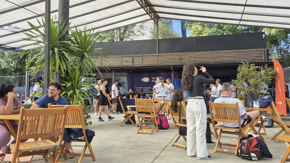 Acessibilidade: Local do evento, há uma tenda branca, com diversas cadeiras de madeira, o ambiente está cheio de participantes e familiares sentados conversando