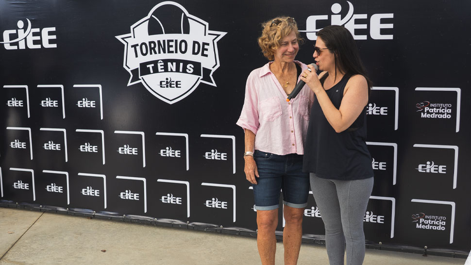 Duas mulheres, uma ao lado da outra, abraçadas, uma veste camisa preta e outra branca. Ambas falam ao microfone. Ao fundo, se encontra o logo do CIEE.