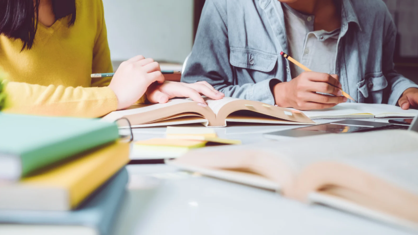 a imagem mostra duas pessoas estudando, com livros na mesa e lápis na mão
