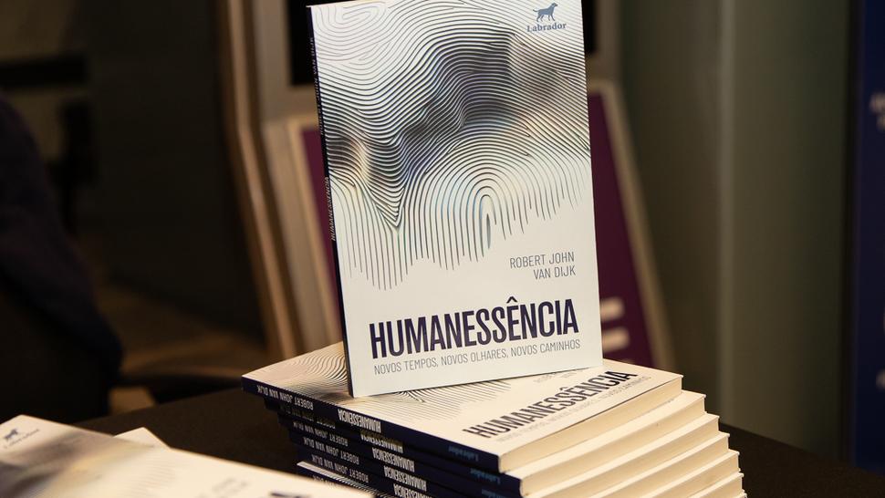 Pilha de livros com a capa em destaque. A obra tem título Humanessência e o nome do autor, Robert John van Dijk. A arte da capa mostra linhas onduladas que formam uma impressão digital estilizada. O fundo é branco com detalhes em azul. Os livros estão sobre uma mesa preta.