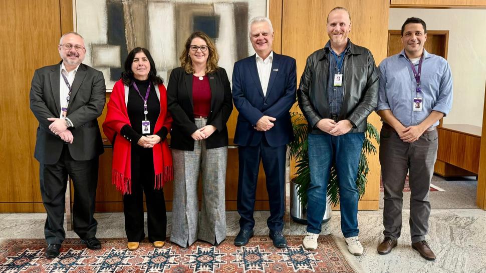 Seis pessoas estão em pé, lado a lado, posando para uma foto em ambiente interno, com parede de madeira e uma obra de arte abstrata ao fundo. Da esquerda para a direita.