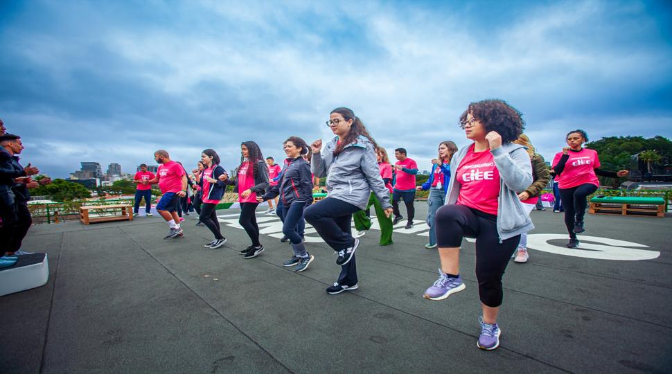Jovens estagiários participam de uma atividade física coletiva ao ar livre. Todos usam camisetas rosa com o logo do CIEE. Eles estão em movimento, levantando uma das pernas como parte do exercício. Ao fundo, o céu está nublado e é possível ver árvores e prédios da cidade.