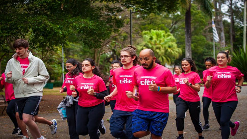 Fotografia de um grupo de pessoas correndo em um parque arborizado. Todos vestem camisetas rosa com a inscrição “Treino Exclusivo CIEE” na frente. Na linha de frente, três homens e três mulheres correm concentrados, com expressões de foco e determinação. Ao fundo, outras pessoas participam da corrida em meio às árvores.