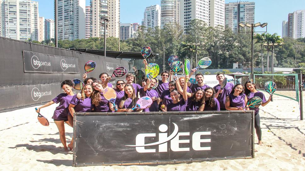 Grupo de jovens estagiários em uma quadra de areia, todos usando camisetas roxas. Eles posam sorridentes atrás de uma placa preta com o logotipo do CIEE em branco, segurando raquetes de beach tênis.