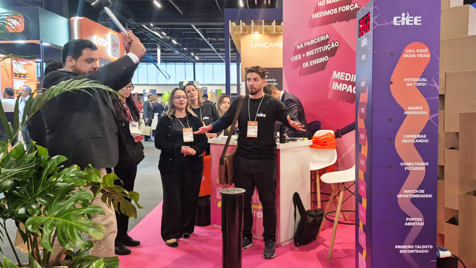 imagem mostra o estande do CIEE trabalhado na cor rosa e roxo. A equipe do CIEE veste roupas pretas e um homem de terno está empunhando um martelo no teste de força.