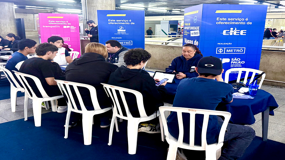 Jovens sendo atendidos em um balcão de orientação do CIEE dentro da estação Sé do Metrô de São Paulo. À mesa, profissionais com coletes azuis e crachás conversam e fazem cadastros em tablets e papéis. Atrás, um painel azul destaca informações sobre vagas de Estágio e Aprendizagem.