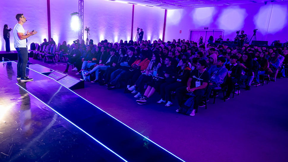 Rodrigo Dib apresentando palestra com camiseta branca em ambiente com iluminação roxa.