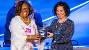 Eunice Prudente segurando o troféu que recebeu orgulhosa em cima do palco. Usa vestes brancas com detalhes florais rosas.