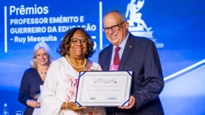 Eunice Prudente segurando o troféu que recebeu orgulhosa em cima do palco. Usa vestes brancas com detalhes florais rosas.