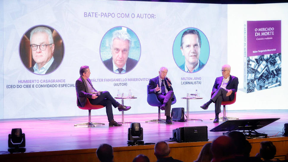 Imagem de um bate-papo no palco do evento de lançamento do livro “O Mercado da Morte: Conexões e Realidades”, realizado no CIEE. Sentados em poltronas, da esquerda para a direita, estão o jornalista Mílton Jung, o autor Wálter Fanganiello Maierovitch e o CEO do CIEE, Humberto Casagrande.