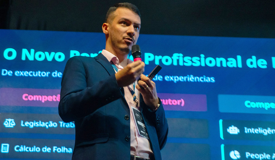 Homem vestido com terno azul-marinho e camisa rosa clara está em pé sobre um palco, segurando um microfone em uma mão e um controle remoto na outra, enquanto apresenta uma palestra. Atrás dele, há um telão com o título “O Novo Perfil do Profissional de RH”.