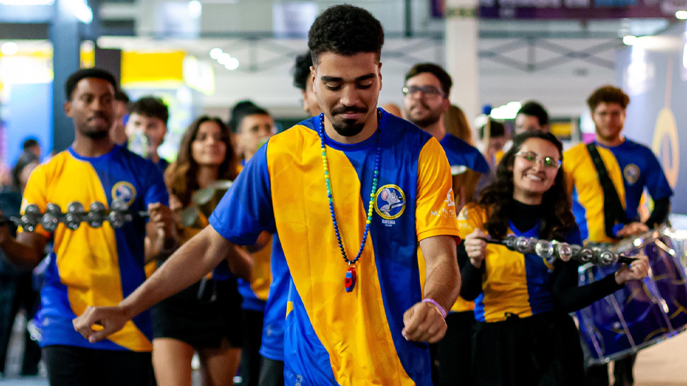 Acessibilidade: Diversos jovens vestindo abadás amarelo e azul tocando instrumentos de baterias de escola de samba.